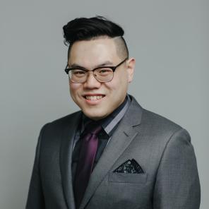 An Asian man with glasses smiles at the camera. He wears a grey suit with a dark striped shirt and a purple tie. His dark hair is cropped short on the sides, and is longer on top, styled to the side.